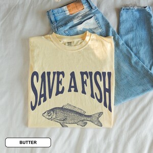 May include: A pale yellow t-shirt featuring the navy blue text "SAVE A FISH" and a fish illustration. Light blue denim jeans are next to the shirt. The shirt is labeled "BUTTER".