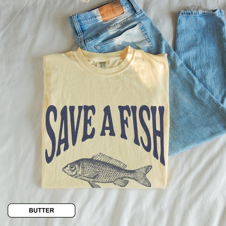 May include: A pale yellow t-shirt featuring the navy blue text "SAVE A FISH" and a fish illustration. Light blue denim jeans are next to the shirt. The shirt is labeled "BUTTER".