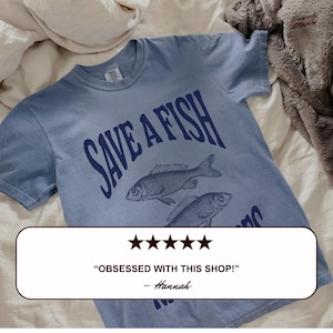 May include: A pale blue t-shirt with the navy blue text "SAVE A FISH" and illustrations of two fish. The shirt also features a customer review that says "OBSESSED WITH THIS SHOP!" and is signed "- Hannah."
