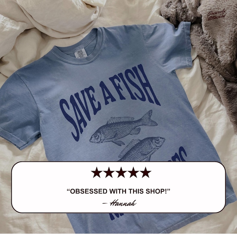 May include: A pale blue t-shirt with the navy blue text "SAVE A FISH" and illustrations of two fish. The shirt also features a customer review that says "OBSESSED WITH THIS SHOP!" and is signed "- Hannah."