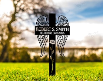 May include: Black metal memorial cross with angel wings and a floral design. The cross features the name "ROBERT S. SMITH" and dates "09.20.1962-06.15.2025". The cross is set in a grassy field with a blurred background of trees and sky.