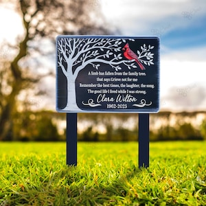 May include: A black memorial garden plaque with a white tree design, a red cardinal, and the text "A limb has fallen from the family tree...Clara Wilton 1962-2025." The plaque is mounted on two black posts and set in green grass.