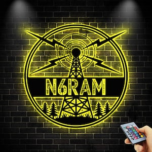 Può includere: Un'insegna illuminata di giallo con la scritta "N6RAM" all'interno di un cerchio. Il design include un'antenna radio, fulmini e alberi, su uno sfondo di muro di mattoni neri. Un telecomando è visibile in basso a destra.