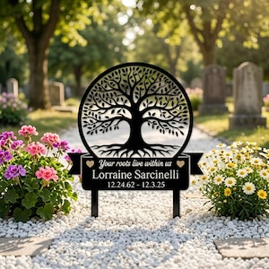 Könnte beinhalten: Schwarzer Metall-Gedenkpfahl mit einem Baum des Lebens-Design in einem Kreis. Der Pfahl trägt den Text "Your roots live within us" und den Namen Lorraine Sarcinelli mit den Daten 12.24.62 - 12.3.25. Das Denkmal befindet sich in einem Garten.