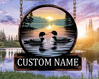 May include: A circular wooden sign depicting a lake scene with two loons, a sunset, and the text "CUSTOM NAME" in white. The sign is hung by chains from a wooden branch.