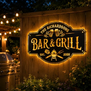 Könnte beinhalten: Ein beleuchtetes Schild in Schwarz und Gold für eine Bar und einen Grill. Auf dem Schild steht "The Richardsons BAR & GRILL EST. 2026" mit Bildern von Bierkrügen, einem Grill und Grillutensilien. Das Schild ist an einem Holzzaun befestigt.