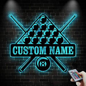May include: A black and blue neon sign featuring a pool rack of billiard balls, two crossed pool cues, and an eight ball. The sign has the text "CUSTOM NAME" in a rectangular banner. The sign is mounted on a brick wall.
