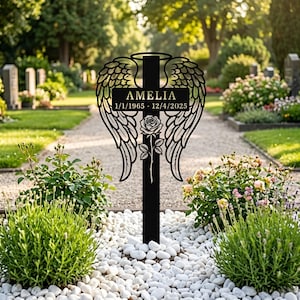 May include: A black metal memorial stake featuring a central cross design adorned with large angel wings on both sides, a halo at the top, and a single rose at the base. The cross displays the name AMELIA and the dates 1/1/1965 - 12/4/2025.