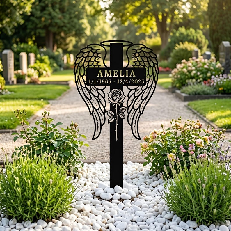 May include: A black metal memorial stake featuring a central cross design adorned with large angel wings on both sides, a halo at the top, and a single rose at the base. The cross displays the name AMELIA and the dates 1/1/1965 - 12/4/2025.