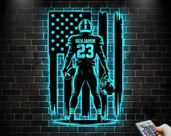 May include: A neon blue LED light featuring a football player in front of an American flag design. The player's jersey displays the name "BENJAMIN" and the number "23". The light is mounted on a dark brick wall.