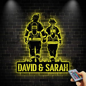 May include: A black and yellow illuminated sign depicting two firefighters holding hands, set against a brick wall. The sign displays the names "DAVID & SARAH" in yellow lettering, with a remote control in the lower right corner.