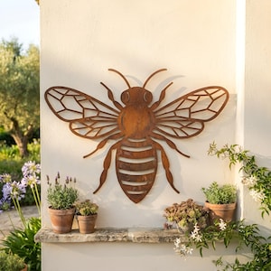 Puede incluir: Una pieza de arte mural de metal color óxido con forma de abeja, que presenta patrones de alas caladas intrincados y un cuerpo segmentado. La abeja está montada en una pared clara y tiene un acabado oxidado y desgastado con una textura visible.