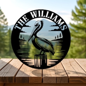 Puede incluir: Arte de pared de metal negro circular con un pelícano posado en un poste de madera, con un paisaje de lago y montañas. El diseño incluye el texto "THE WILLIAMS". Ideal para decoración exterior o costera.
