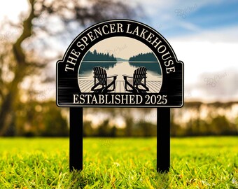 May include: A black and white sign for "The Spencer Lakehouse," established in 2025. The sign features a lake scene with two Adirondack chairs on a dock. The sign is mounted on two black posts and set in a grassy area.