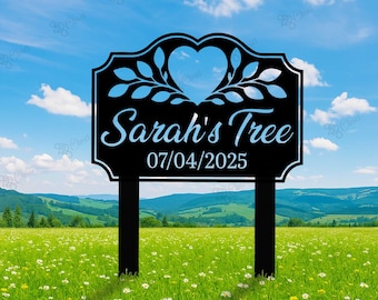 May include: Black metal garden sign with the text "Sarah's Tree" and the date 07/04/2025. The sign has a heart-shaped cutout and leaf design. It is placed in a field of green grass with a blue sky background.