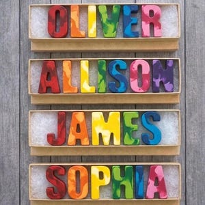 May include: Four boxes, each with a name spelled out in crayon letters. The names are Oliver, Allison, James, and Sophia. The crayons are in various colours, including red, orange, yellow, green, blue, and purple. The boxes are on a grey wooden background.