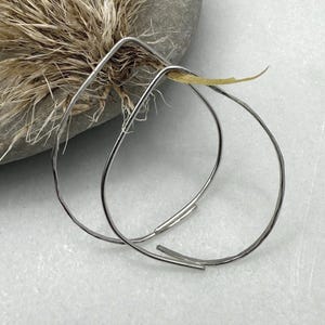May include: A pair of silver-toned hoop earrings with a hammered texture. The earrings have a teardrop shape and a minimalist design. The background includes a gray stone and dried grasses, creating a natural aesthetic.