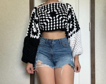 May include: Crocheted crop top in a black and white geometric pattern. The top features long sleeves with a color block design, transitioning from black to white. Paired with denim shorts.