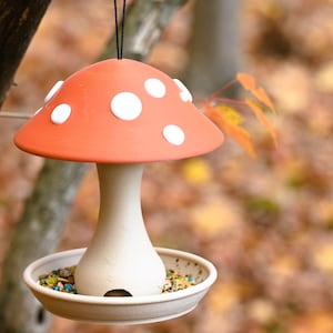 May include: A hanging bird feeder shaped like a mushroom with a terracotta-colored cap featuring white circular spots. The stem and base are a light beige color, and the base contains a mixture of birdseed. It hangs from a thin black cord.