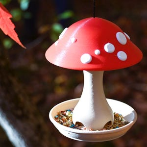 May include: A hanging bird feeder shaped like a mushroom with a bright red cap featuring white spots. The stem is off-white and supports a circular base filled with birdseed. The feeder is suspended by a thin black string from above.