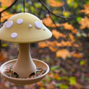 May include: A hanging bird feeder shaped like a mushroom with a pale yellow cap featuring white circular spots. The base is a smooth, light tan stem connected to a circular tray filled with colorful birdseed, suspended by a thin black cord.