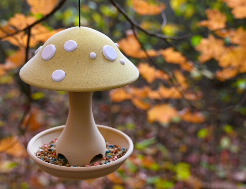 May include: A hanging bird feeder shaped like a mushroom with a pale yellow cap featuring white circular spots. The base is a smooth, light tan stem connected to a circular tray filled with colorful birdseed, suspended by a thin black cord.