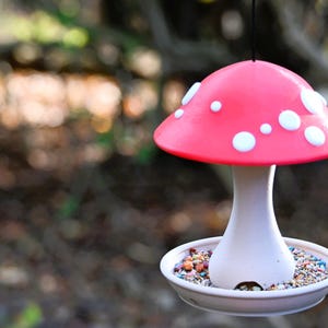 May include: A hanging bird feeder shaped like a mushroom with a bright pink cap featuring white circular spots. The stem is white and connects to a circular base filled with small, multi-colored birdseed. A thin black cord hangs from the top.