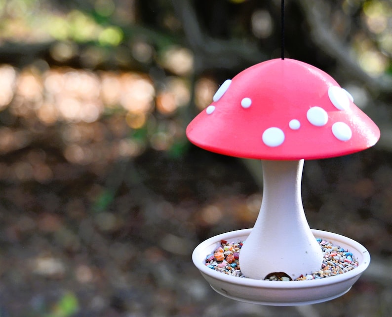 May include: A hanging bird feeder shaped like a mushroom with a bright pink cap featuring white circular spots. The stem is white and connects to a circular base filled with small, multi-colored birdseed. A thin black cord hangs from the top.