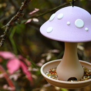 May include: A hanging bird feeder shaped like a mushroom with a textured lavender cap featuring white circular spots. The stem is a smooth light beige color, and the base is a wide, shallow tray filled with a variety of small bird seeds.