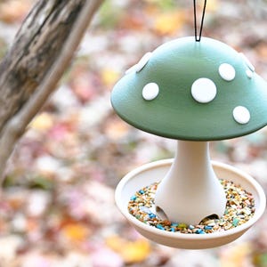 May include: A mushroom-shaped bird feeder featuring a matte green cap with white circular spots. The base is a light beige color with a circular tray at the bottom filled with a variety of colorful bird seeds. A black string is attached to the top.