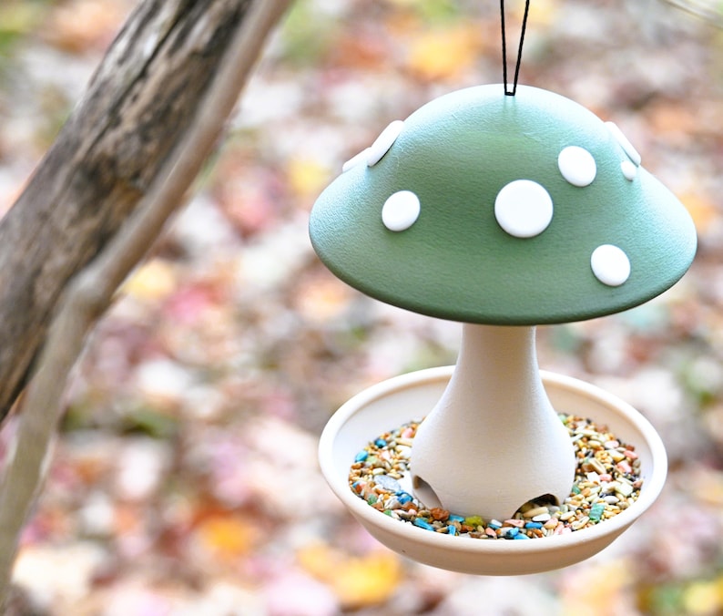 May include: A mushroom-shaped bird feeder featuring a matte green cap with white circular spots. The base is a light beige color with a circular tray at the bottom filled with a variety of colorful bird seeds. A black string is attached to the top.