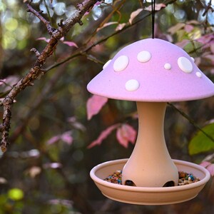 May include: A hanging bird feeder shaped like a mushroom with a light purple cap featuring white circular spots. The stem and base are a neutral tan color, and the base contains a mixture of birdseed. A black string is attached to the top.
