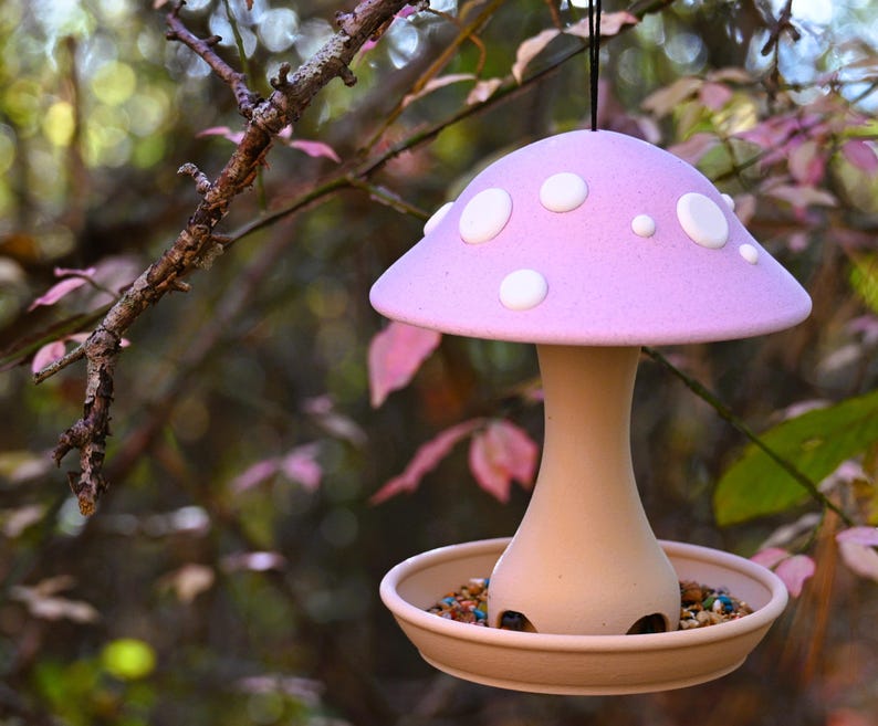 May include: A hanging bird feeder shaped like a mushroom with a light purple cap featuring white circular spots. The stem and base are a neutral tan color, and the base contains a mixture of birdseed. A black string is attached to the top.