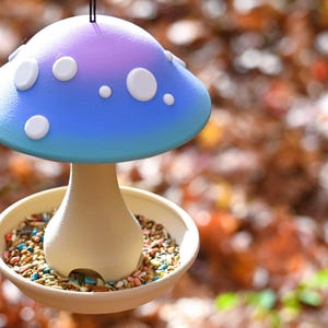 May include: A mushroom-shaped bird feeder featuring a blue and purple gradient cap with white circular spots. The stem is a light beige color and sits in the center of a circular tray filled with a variety of small bird seeds and grains.