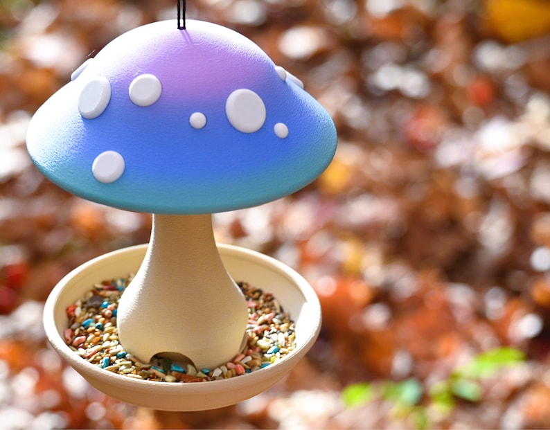 May include: A mushroom-shaped bird feeder featuring a blue and purple gradient cap with white circular spots. The stem is a light beige color and sits in the center of a circular tray filled with a variety of small bird seeds and grains.