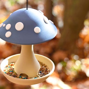 May include: A hanging bird feeder shaped like a mushroom with a matte blue cap featuring raised white spots. The stem is a light beige color with arched openings at the base, resting in a shallow beige tray filled with a mix of bird seeds.