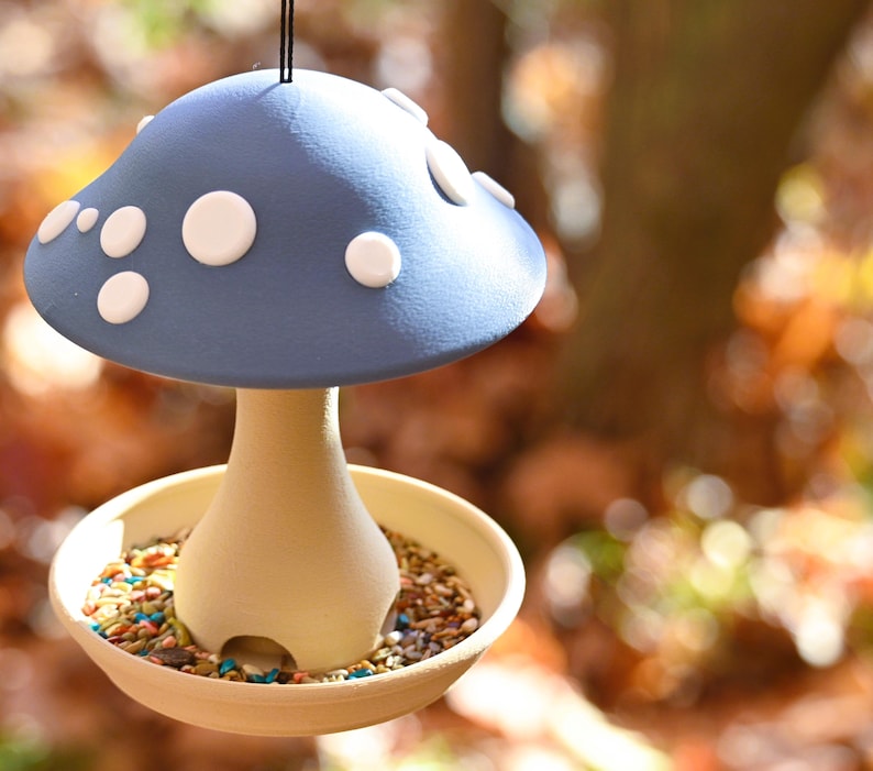 May include: A hanging bird feeder shaped like a mushroom with a matte blue cap featuring raised white spots. The stem is a light beige color with arched openings at the base, resting in a shallow beige tray filled with a mix of bird seeds.