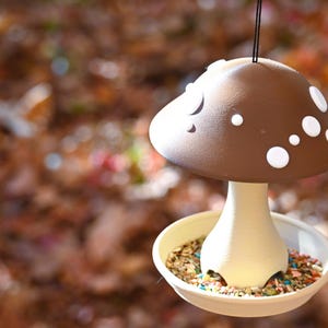 May include: A hanging bird feeder shaped like a mushroom with a brown cap featuring white circular spots. The stem is cream-colored and connects to a circular base tray filled with a variety of bird seeds, including small colorful pieces.