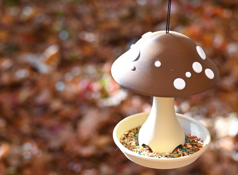 May include: A hanging bird feeder shaped like a mushroom with a brown cap featuring white circular spots. The stem is cream-colored and connects to a circular base tray filled with a variety of bird seeds, including small colorful pieces.