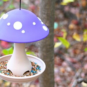 May include: A hanging bird feeder shaped like a mushroom with a purple cap featuring white circular spots. The stem is light beige and connects to a circular base tray filled with multicolored birdseed, suspended by a thin black cord.