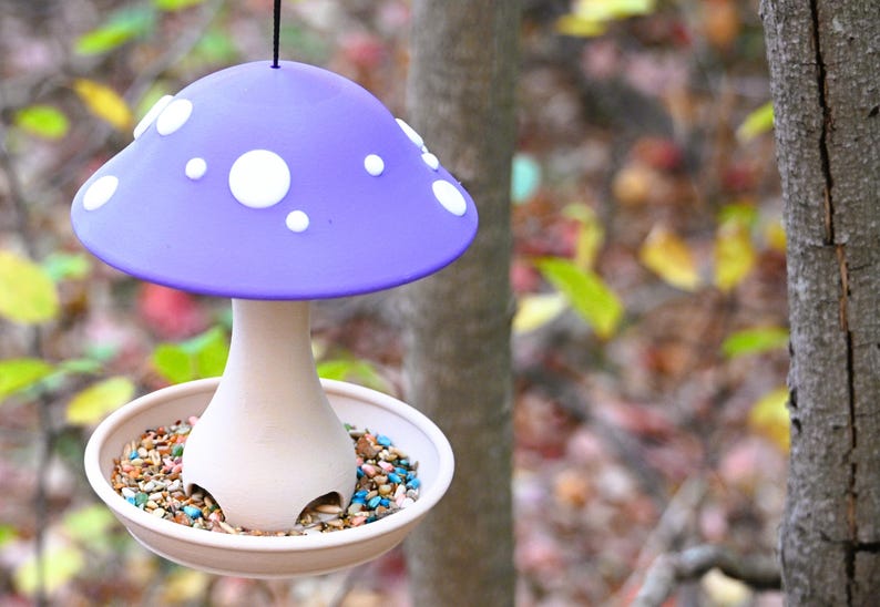 May include: A hanging bird feeder shaped like a mushroom with a purple cap featuring white circular spots. The stem is light beige and connects to a circular base tray filled with multicolored birdseed, suspended by a thin black cord.