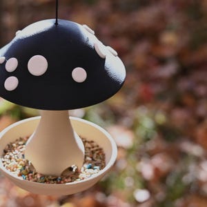 May include: A hanging bird feeder shaped like a mushroom with a dark blue cap featuring white circular spots. The base is a light beige bowl filled with mixed birdseed, surrounding a central stem that supports the wide, overhanging cap.