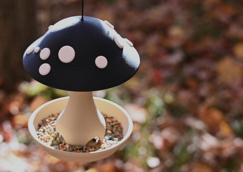 May include: A hanging bird feeder shaped like a mushroom with a dark blue cap featuring white circular spots. The base is a light beige bowl filled with mixed birdseed, surrounding a central stem that supports the wide, overhanging cap.