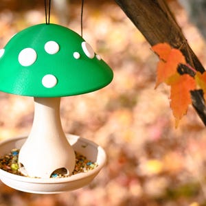 May include: A hanging bird feeder shaped like a mushroom with a bright green cap featuring white circular spots. The stem is a light beige color with arched openings at the base, resting inside a shallow circular tray filled with birdseed.