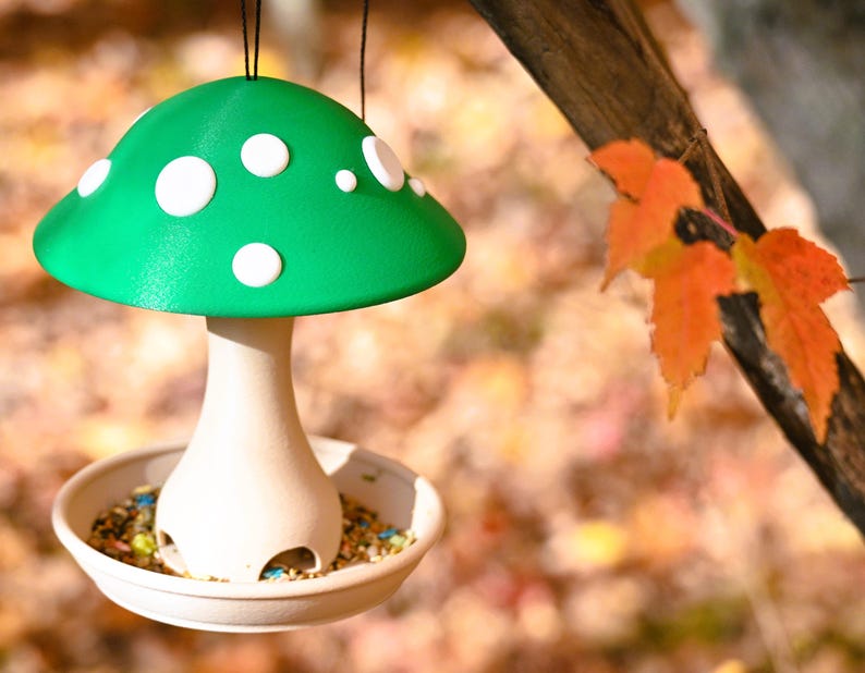 May include: A hanging bird feeder shaped like a mushroom with a bright green cap featuring white circular spots. The stem is a light beige color with arched openings at the base, resting inside a shallow circular tray filled with birdseed.