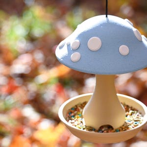 May include: A hanging bird feeder shaped like a mushroom with a light blue cap featuring white circular spots. The base is a cream-colored stem that connects to a circular tray filled with a variety of small, colorful bird seeds and grains.