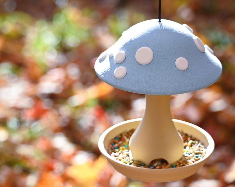 May include: A hanging bird feeder shaped like a mushroom with a light blue cap featuring white circular spots. The base is a cream-colored stem that connects to a circular tray filled with a variety of small, colorful bird seeds and grains.
