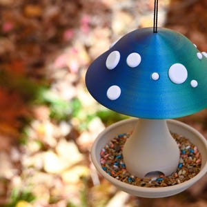 May include: A mushroom-shaped bird feeder featuring a gradient blue and green cap adorned with white circular spots. The base is a light beige, bowl-shaped tray filled with a variety of bird seeds, suspended by a thin black cord for hanging.