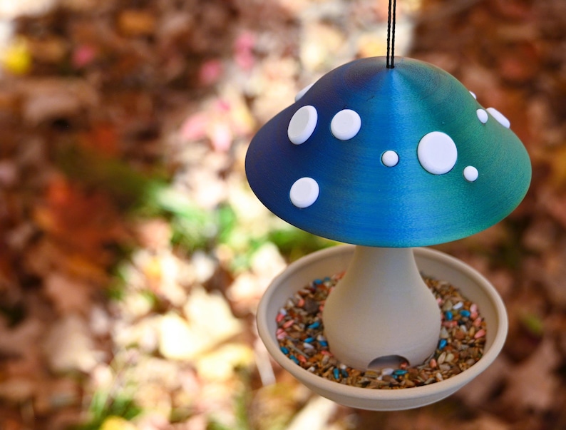 May include: A mushroom-shaped bird feeder featuring a gradient blue and green cap adorned with white circular spots. The base is a light beige, bowl-shaped tray filled with a variety of bird seeds, suspended by a thin black cord for hanging.