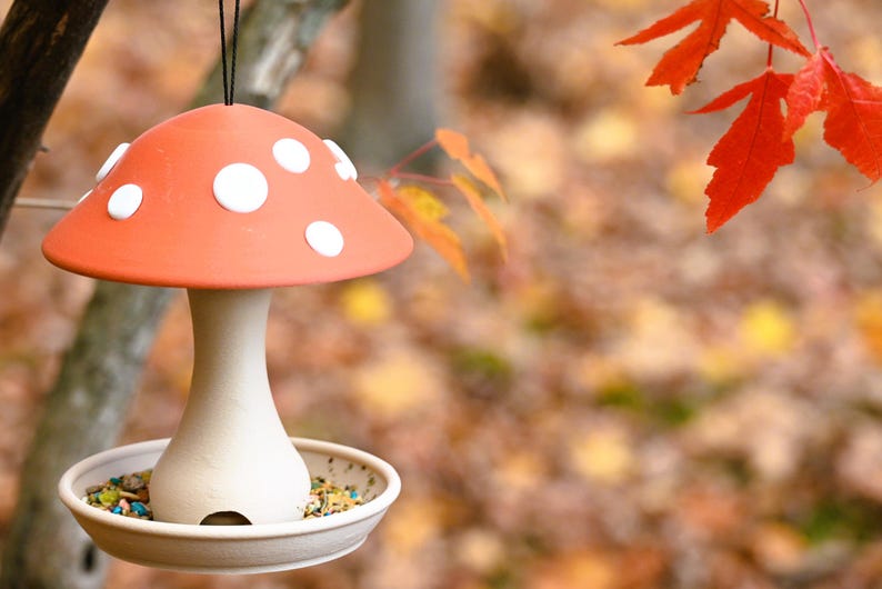 May include: A hanging bird feeder shaped like a mushroom with a terracotta-colored cap featuring white circular spots. The stem and base are a light beige color, and the base contains a mixture of birdseed. It hangs from a thin black cord.